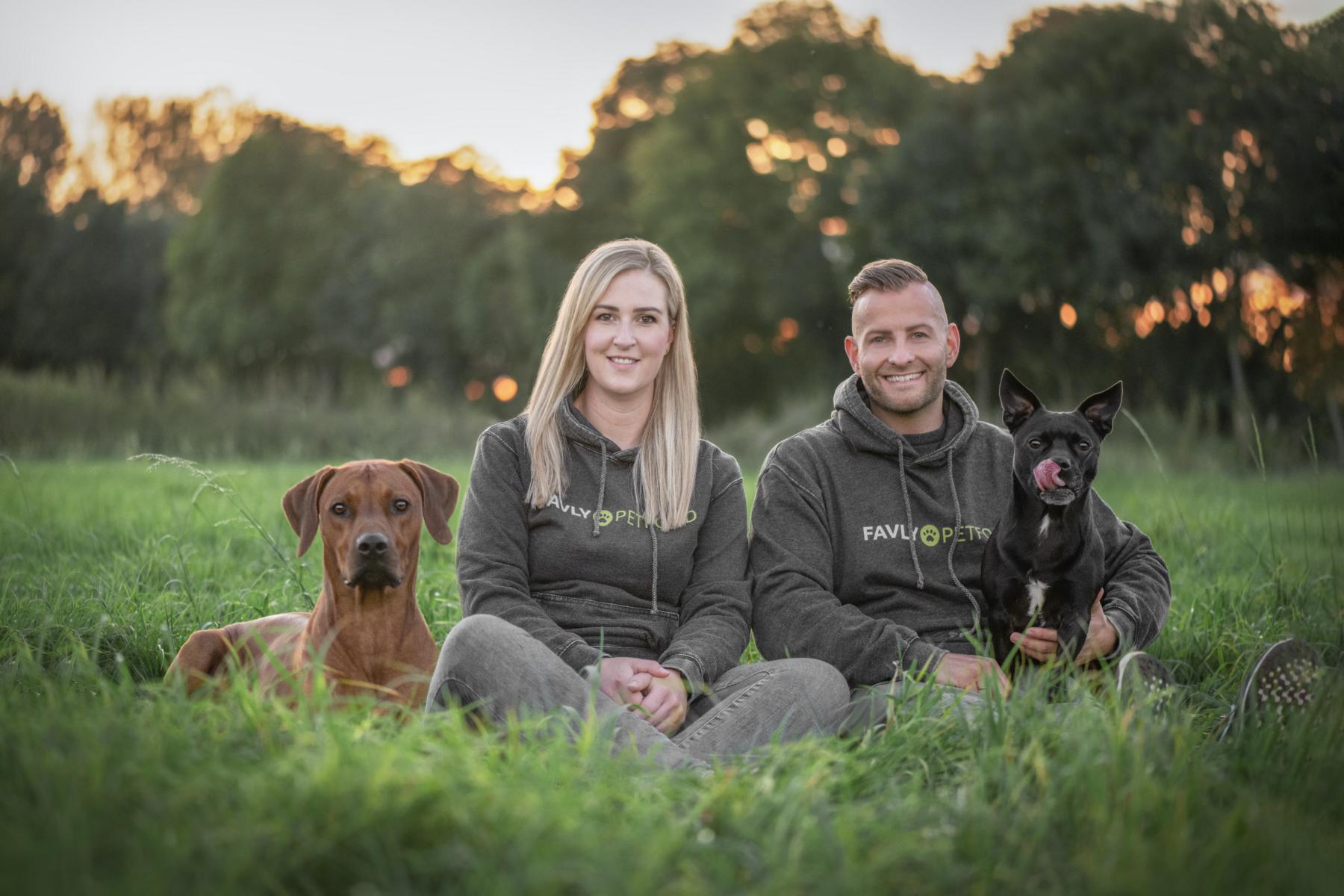 Ein Paar sitzt mit zwei Hunden auf einem Feld und lächelt bei Sonnenuntergang.